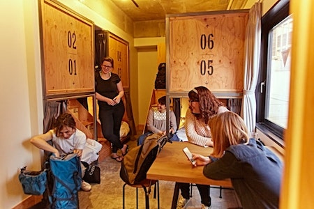 Guests sitting at a table beside numbered wooden capsule beds at Wise Owl Hostels Shibuya