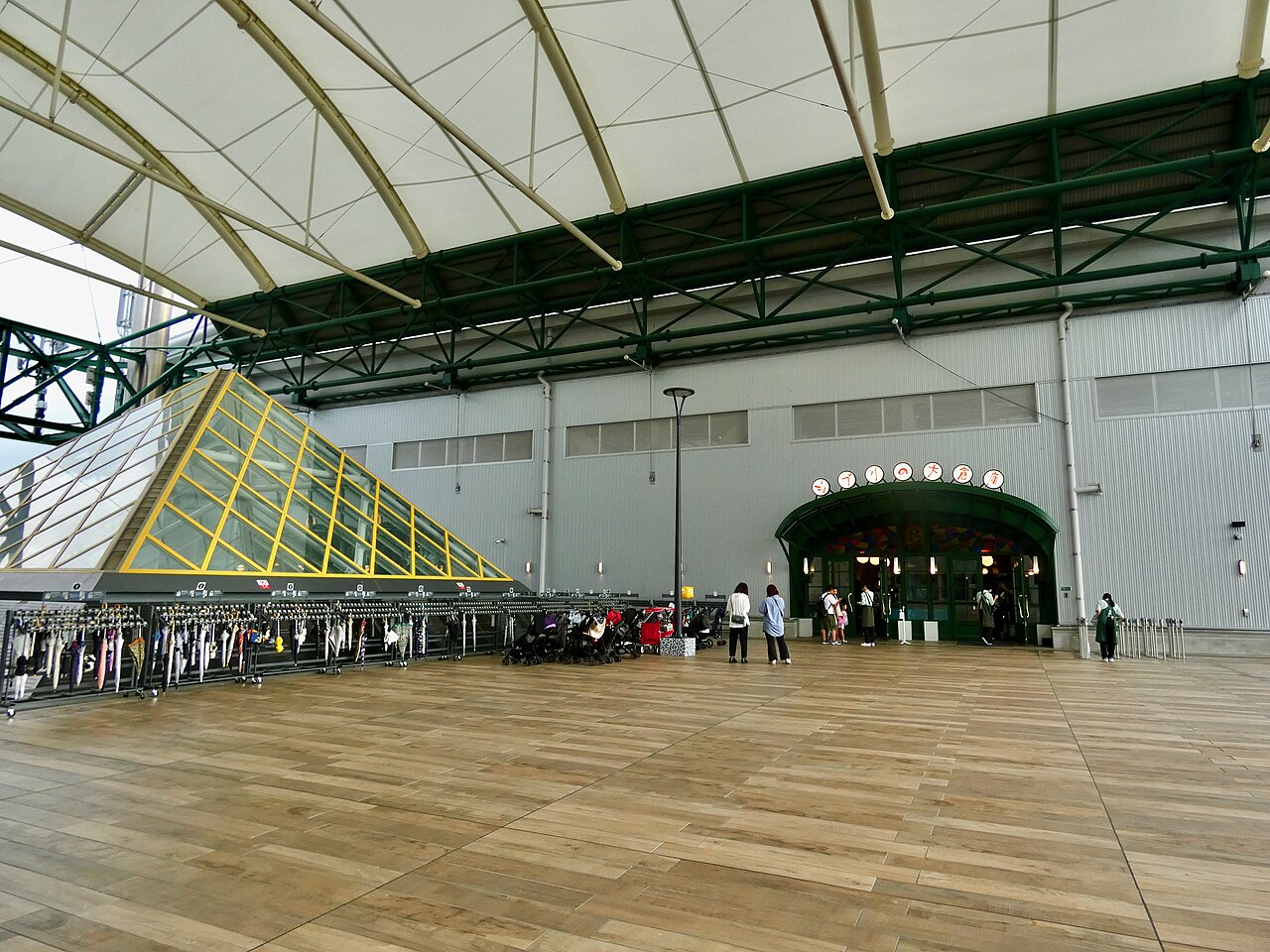The arched green entrance of Ghibli’s Grand Warehouse under a white canopy roof, with visitors queuing on a wooden deck