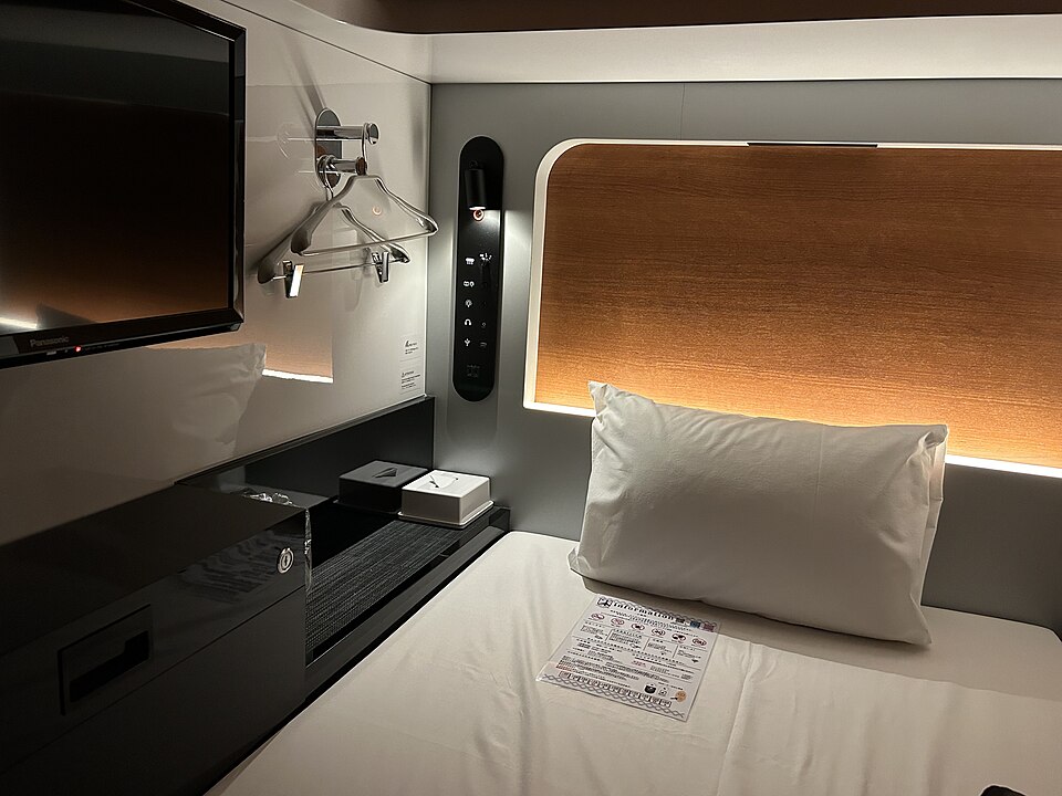 Inside a First Cabin capsule hotel room in Ichigaya, Tokyo, with a single bed, wall mounted TV and wood panelling