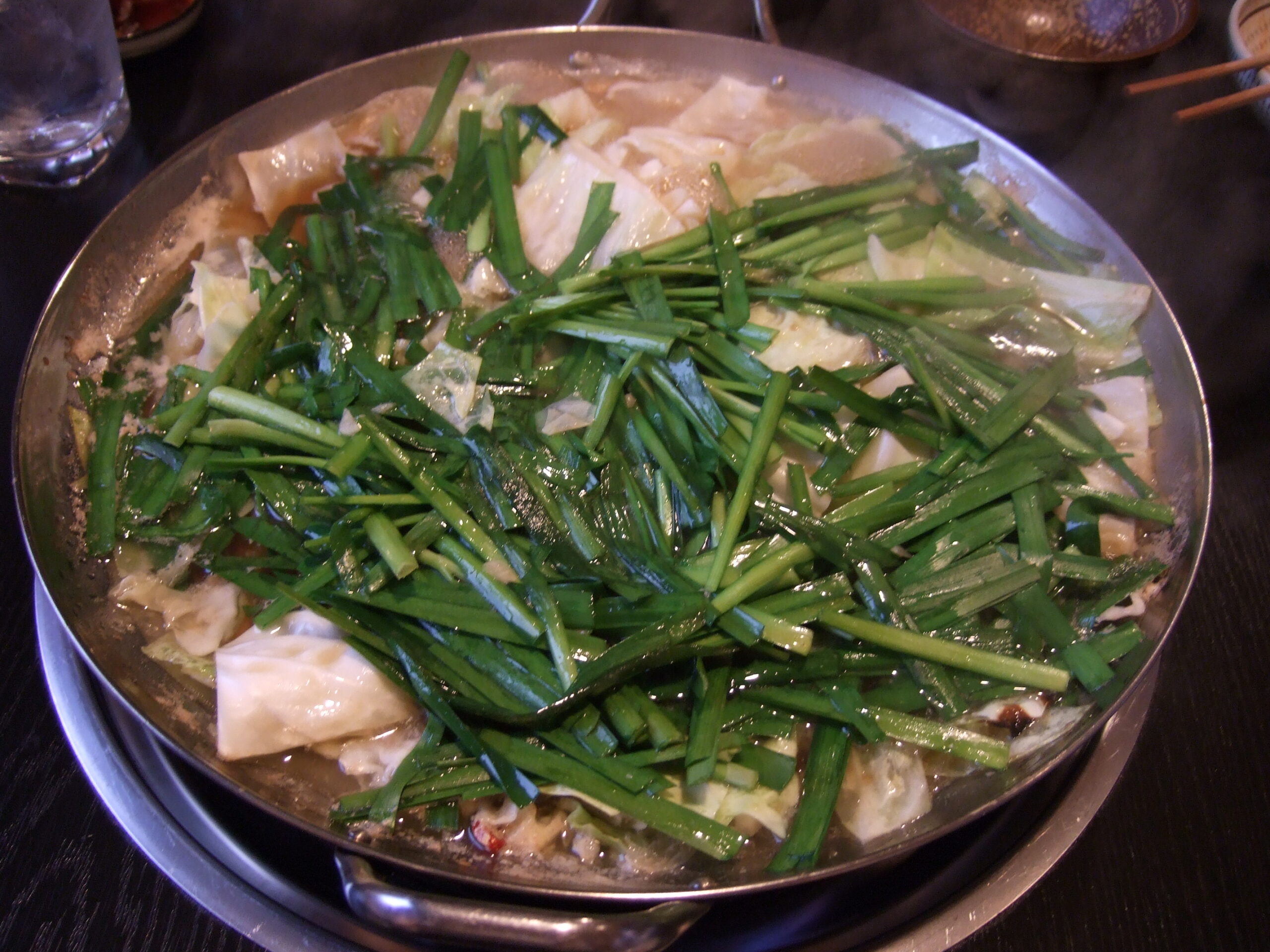 Shallow metal pot of motsunabe topped with a thick layer of garlic chives over cabbage
