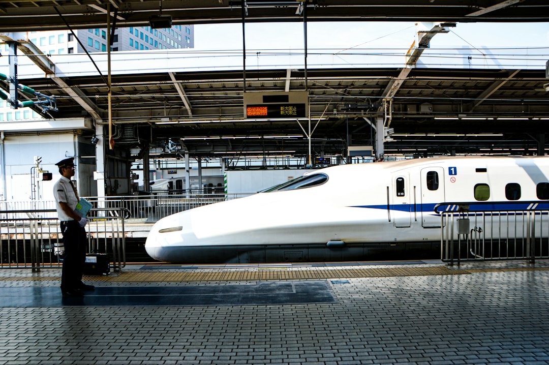 A white Shinkansen bullet train with a blue stripe waiting at a station platform while a member of station staff stands on the platform