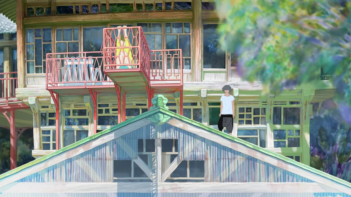 A young man stands on the roof of an old wooden building with red balconies and rows of windows, framed by blurred green leaves.