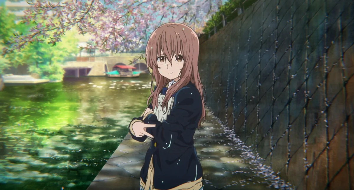 Shoko, with long brown hair and a school blazer, stands on a path beside a green canal as cherry blossom petals drift onto the water.