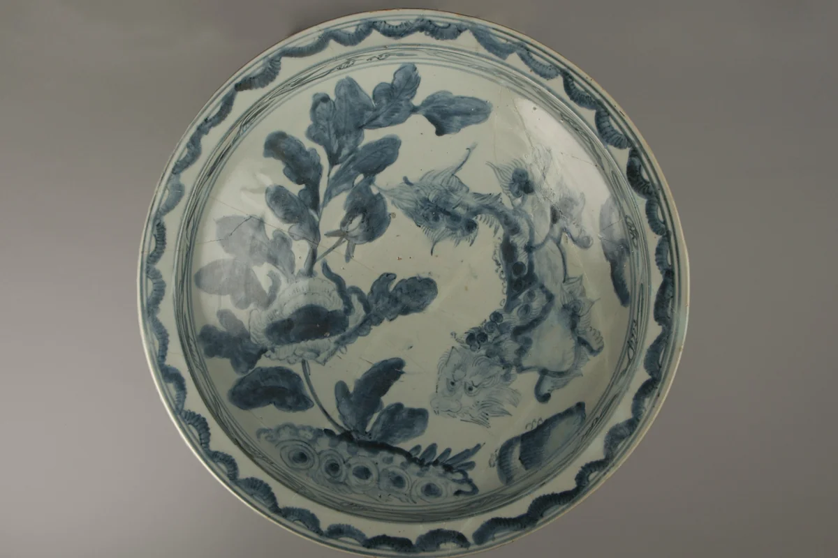 A large round porcelain dish painted in cobalt blue on white, showing a lion among peony leaves inside a border of flowers