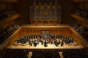 Bach Collegium Japan's choir and period instrument orchestra on stage in a wood panelled concert hall beneath a pipe organ, with a full audience