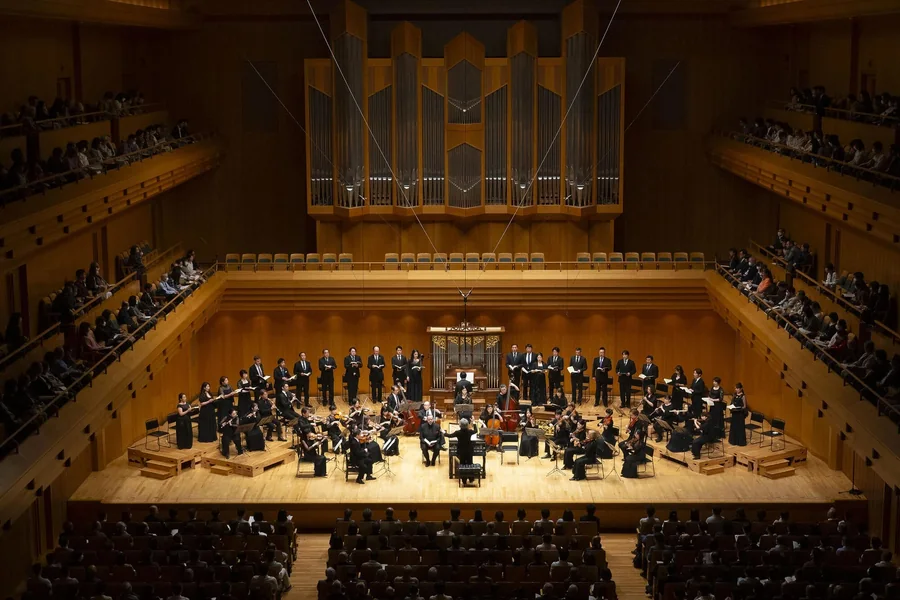 Bach Collegium Japan's choir and period instrument orchestra on stage in a wood panelled concert hall beneath a pipe organ, with a full audience