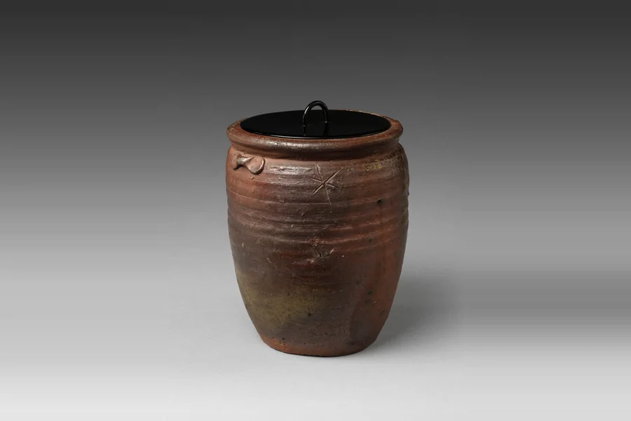A brown unglazed Bizen stoneware water jar with ridged sides, two small lug handles and a black lacquer lid with a ring handle