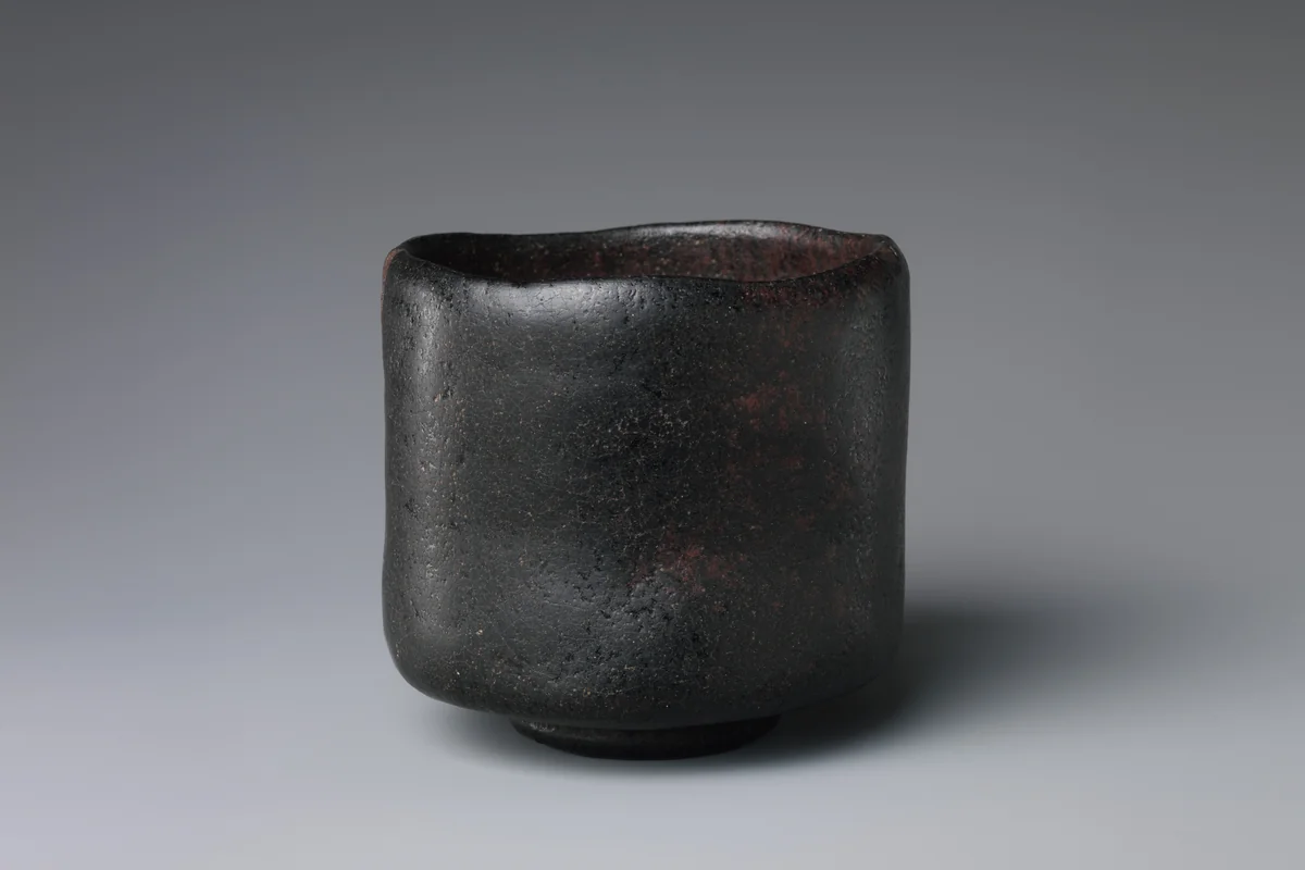A black Raku tea bowl with tall, slightly uneven walls and a matt black glaze, photographed against a grey background