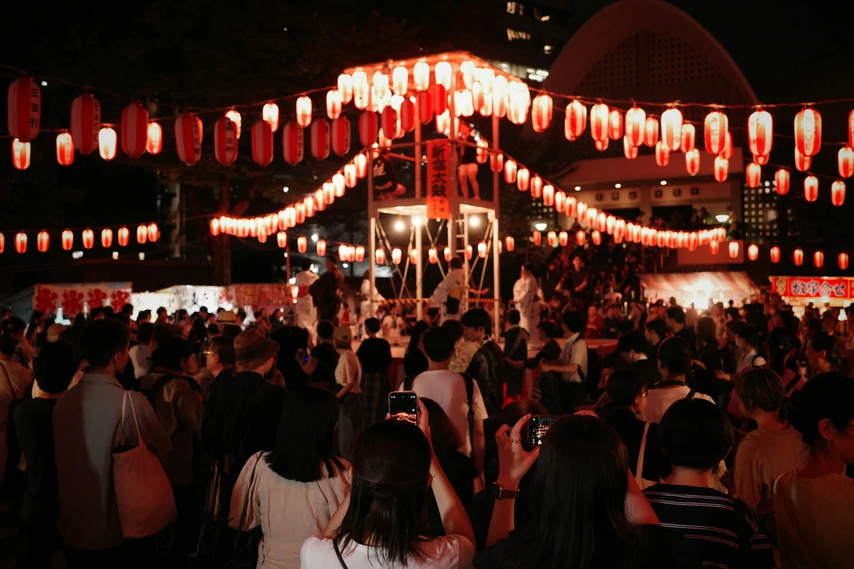 Strings of red and white paper lanterns radiating from a central yagura tower over a crowd at a night summer festival in Japan