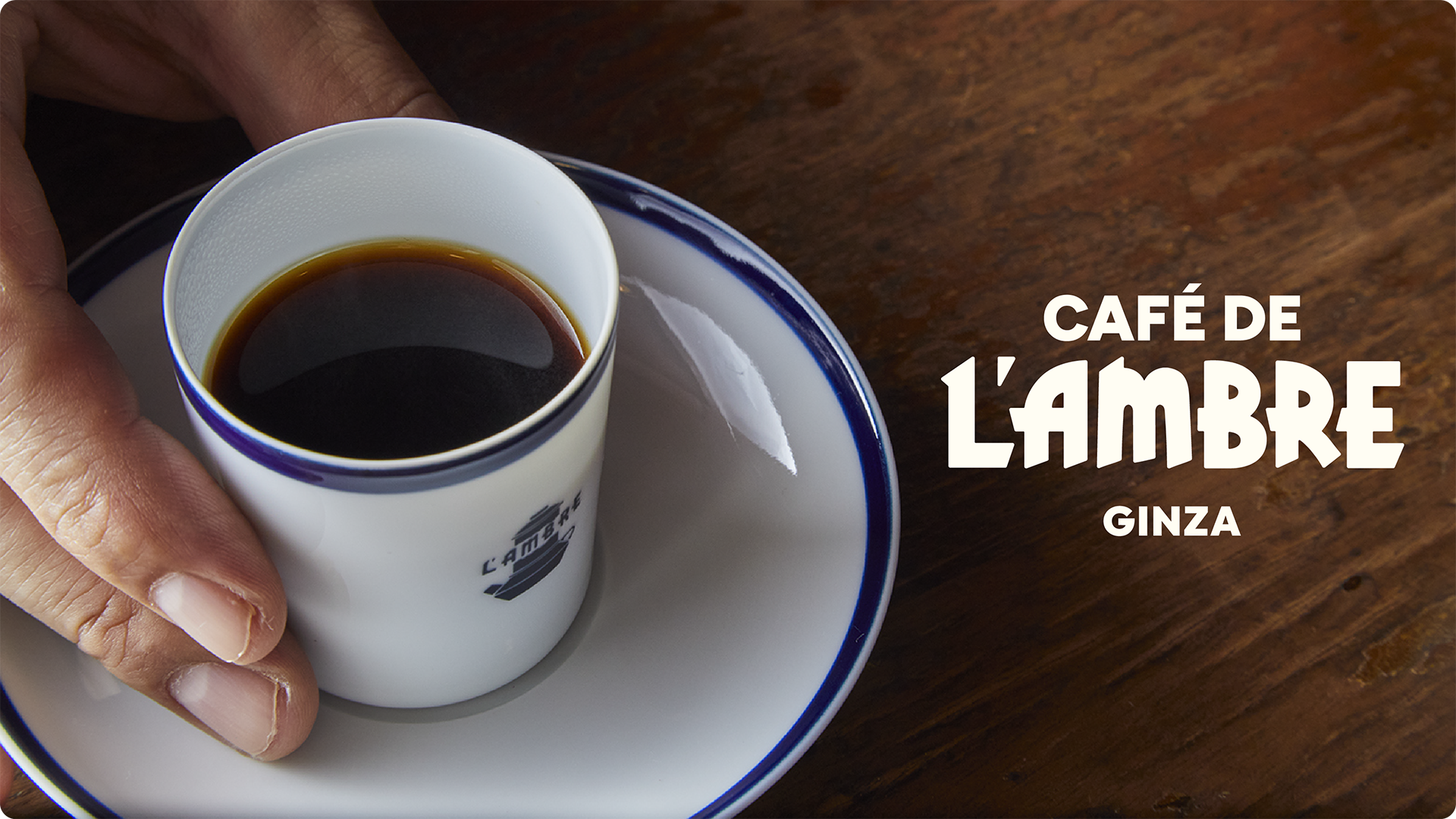 A hand holding a white cup of black coffee on a saucer beside the Café de L'Ambre Ginza logo
