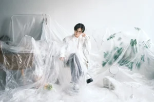 Centimillimental in a white suit and glasses, sitting among sheets of clear plastic in a white room