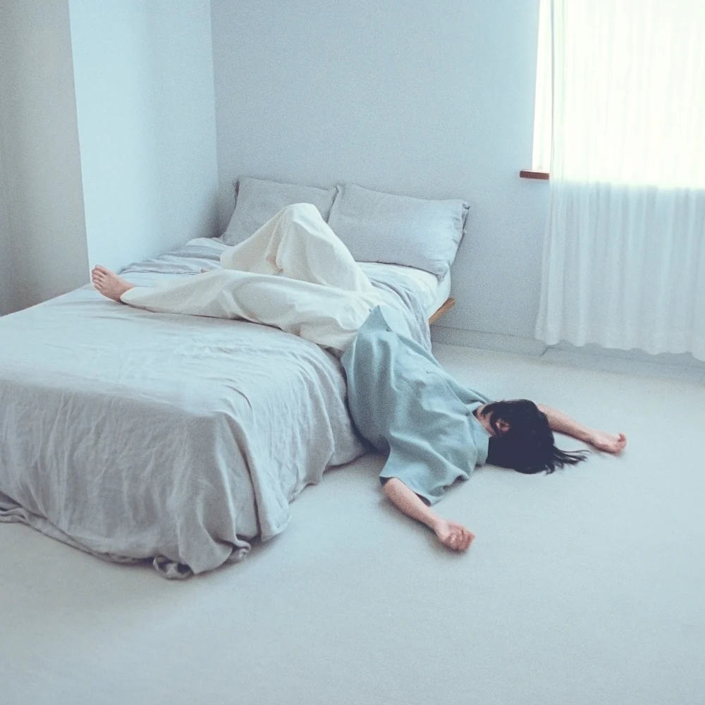 A figure in a pale blue shirt lying face down, half on a bed and half on the floor of a bright white room