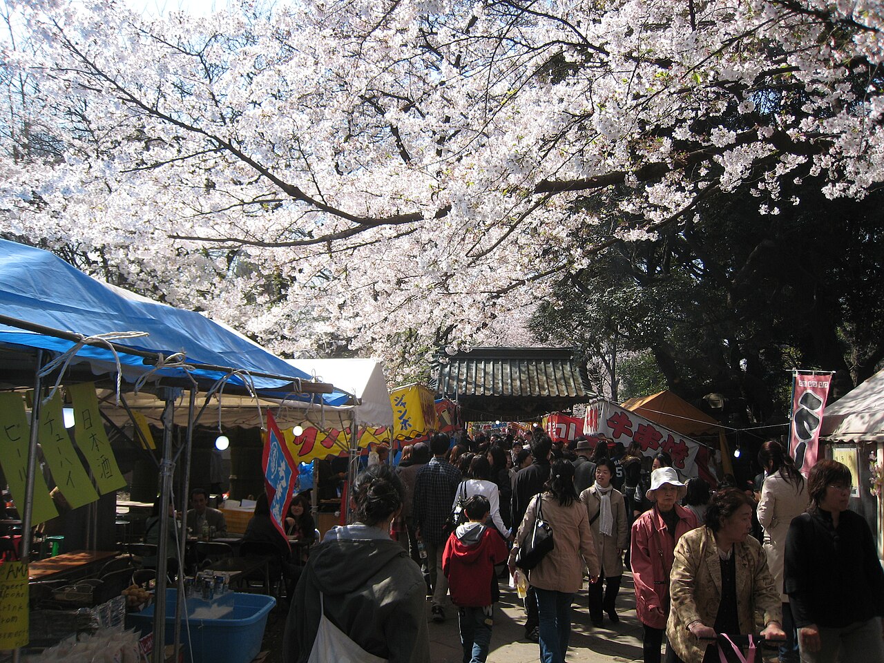 A crowded footpath lined with food stalls under cherry blossoms in Ueno Park, Tokyo