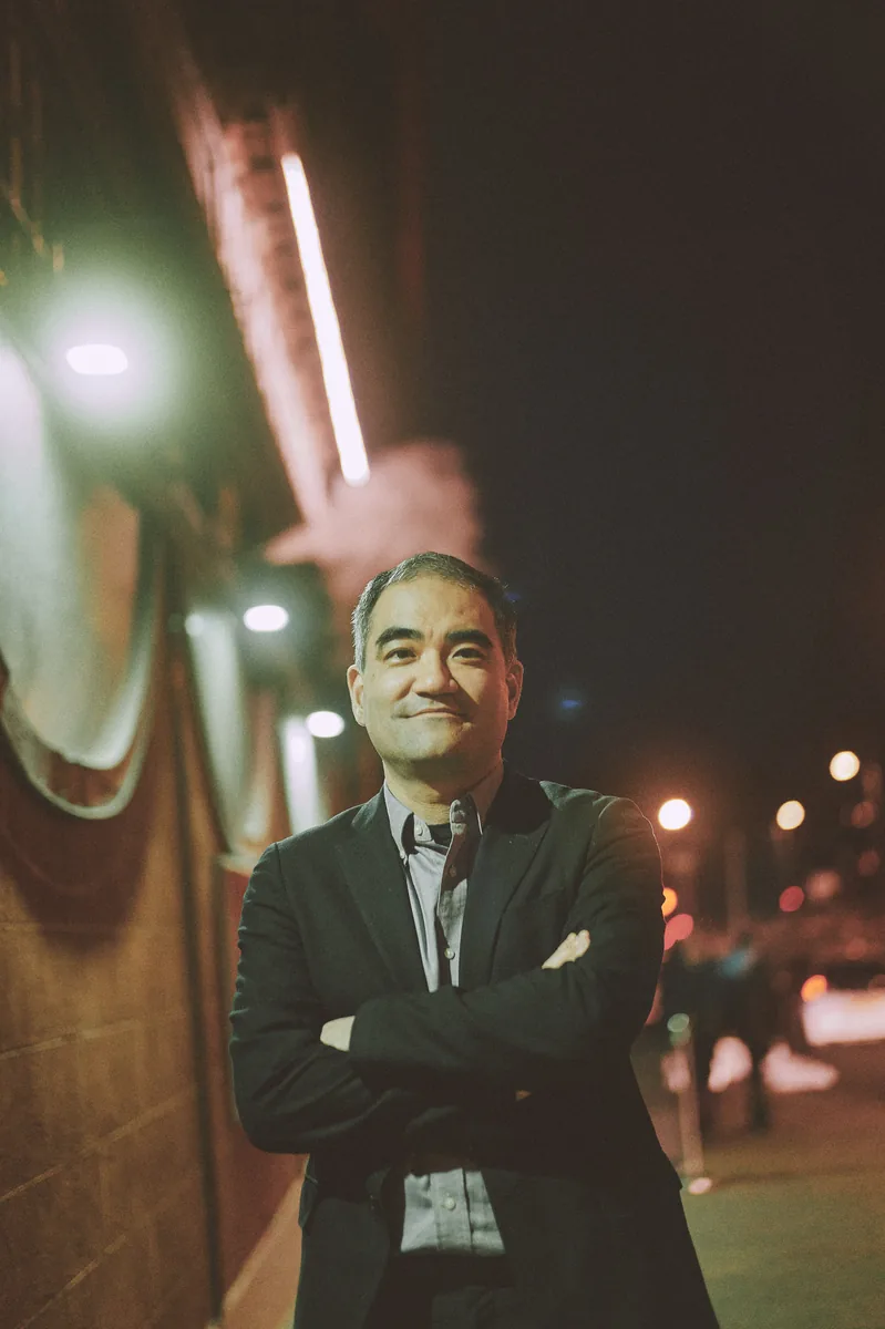 Dai Fujikura at night with his arms folded, standing on a city pavement beside a lit building, street lights behind him.