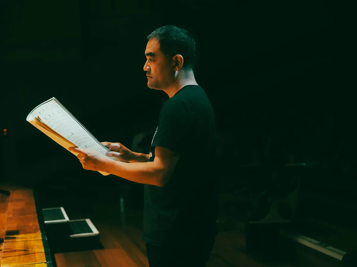 Dai Fujikura standing on a darkened stage, reading from a spiral bound score.