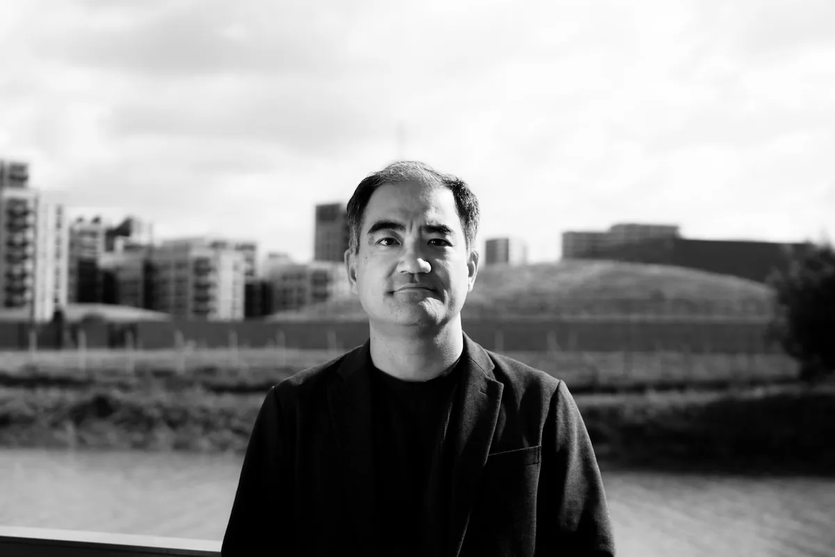 Black and white portrait of Dai Fujikura in a dark jacket, standing by a river with buildings behind him.