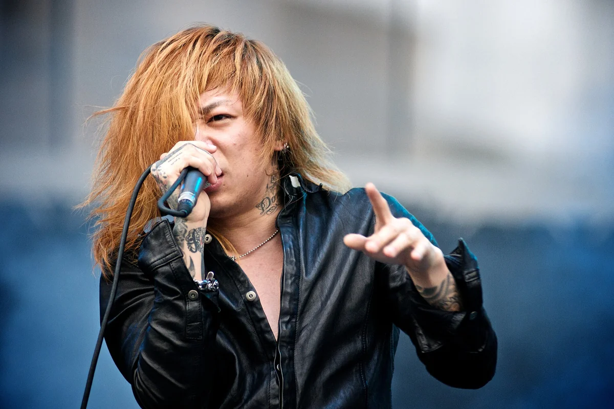 Dir en grey's singer Kyo, with long blond hair and a black leather jacket, singing into a microphone on an outdoor stage