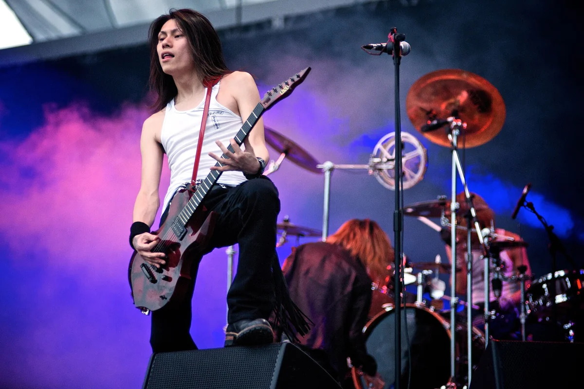 A Dir en grey guitarist in a white vest playing a dark electric guitar on stage, one foot on a monitor, with a drummer behind him in purple stage light