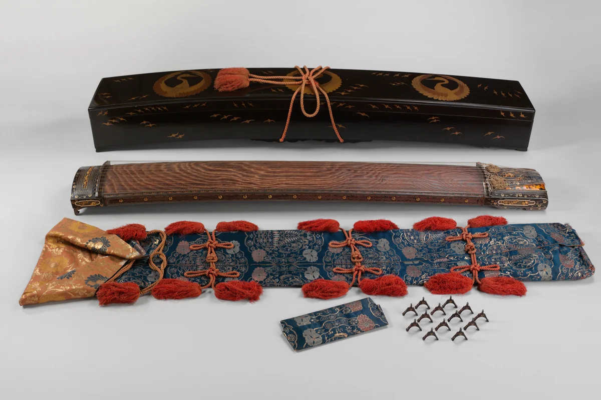 An early 17th century Japanese koto shown with its black lacquer storage case decorated with gold cranes, a blue and gold brocade cover with red tassels, and a small pile of movable bridges
