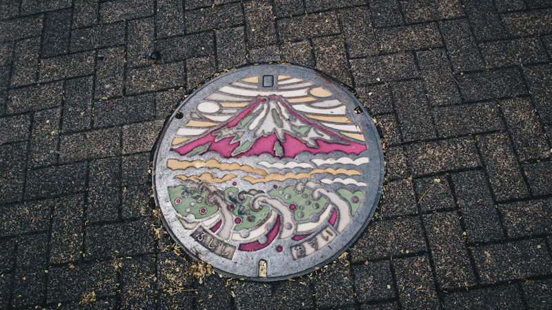 Colour painted manhole cover in Fuji City, Shizuoka, showing Mount Fuji above rolling waves, set into a dark brick pavement