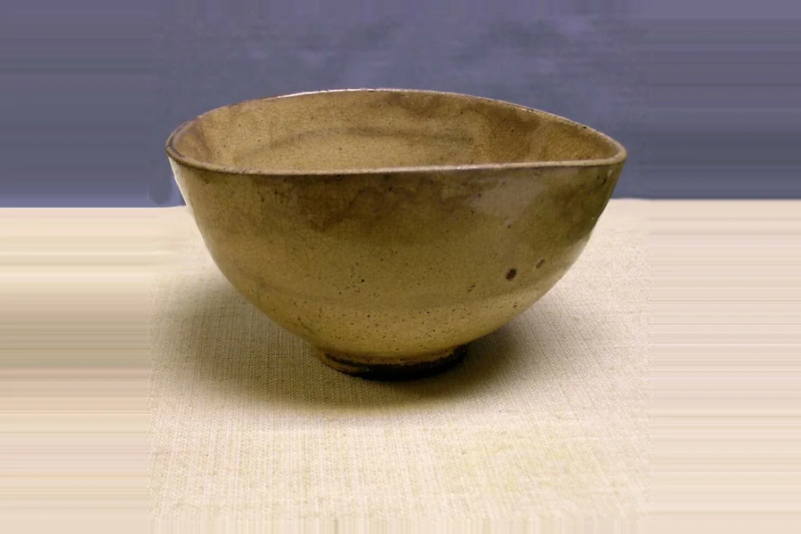 A wide, slightly oval stoneware bowl with a pale olive brown glaze covered in fine crackle, photographed on a cloth