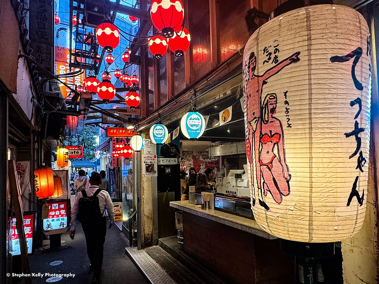 A man walking down a narrow lane in Harmonica Yokocho, Kichijoji, under red paper lanterns beside a yakitori counter