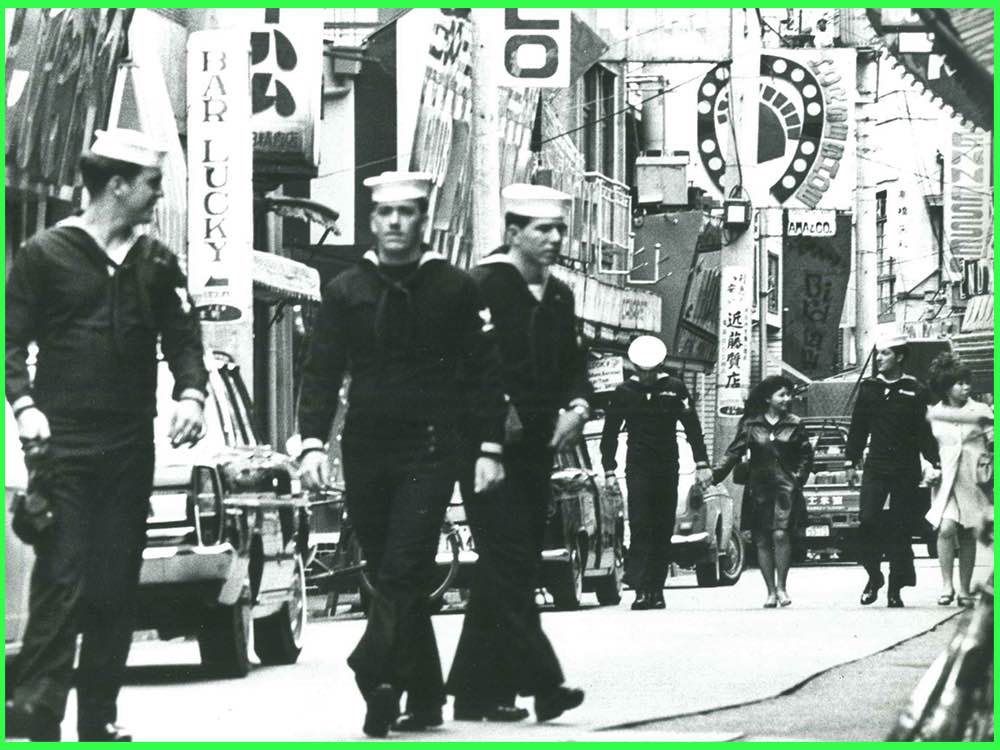Black and white image from History of Postwar Japan as Told by a Bar Hostess (1970): American sailors walk down a street lined with bar signs