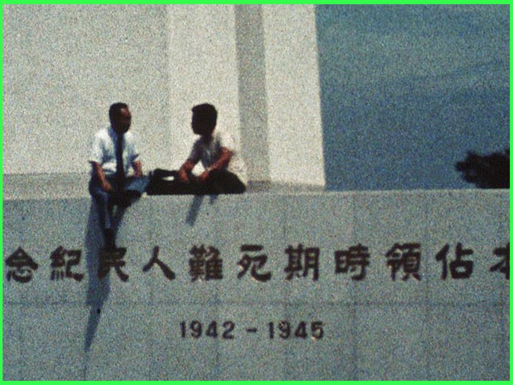 Colour still from In Search of the Unreturned Soldiers: two men sit on top of a war memorial carved with the years 1942 to 1945