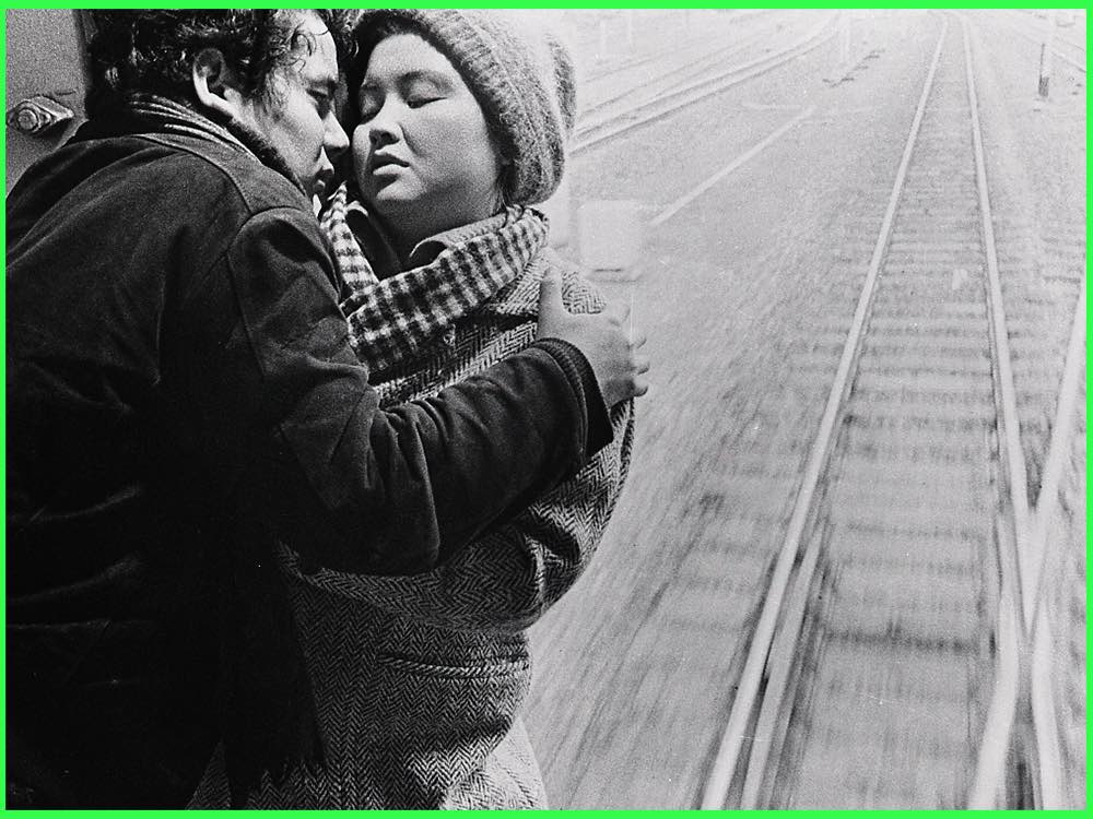 Black and white still from Intentions of Murder (1964): a man in a heavy coat holds a woman in a woollen hat beside a railway track