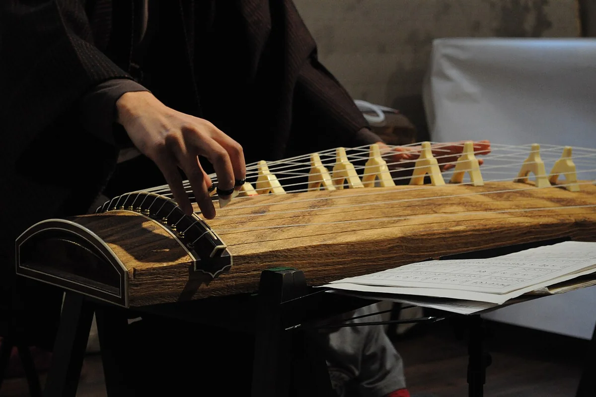 A koto player's right hand plucks the strings of a wooden koto, with a row of white movable bridges standing on the soundboard and sheet music beside it