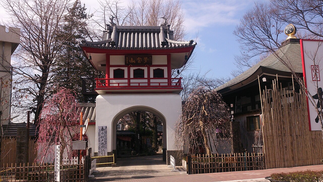 The white and red bell gate of Jogan-ji temple in Nakano, Tokyo, with a pink weeping plum tree in blossom beside it
