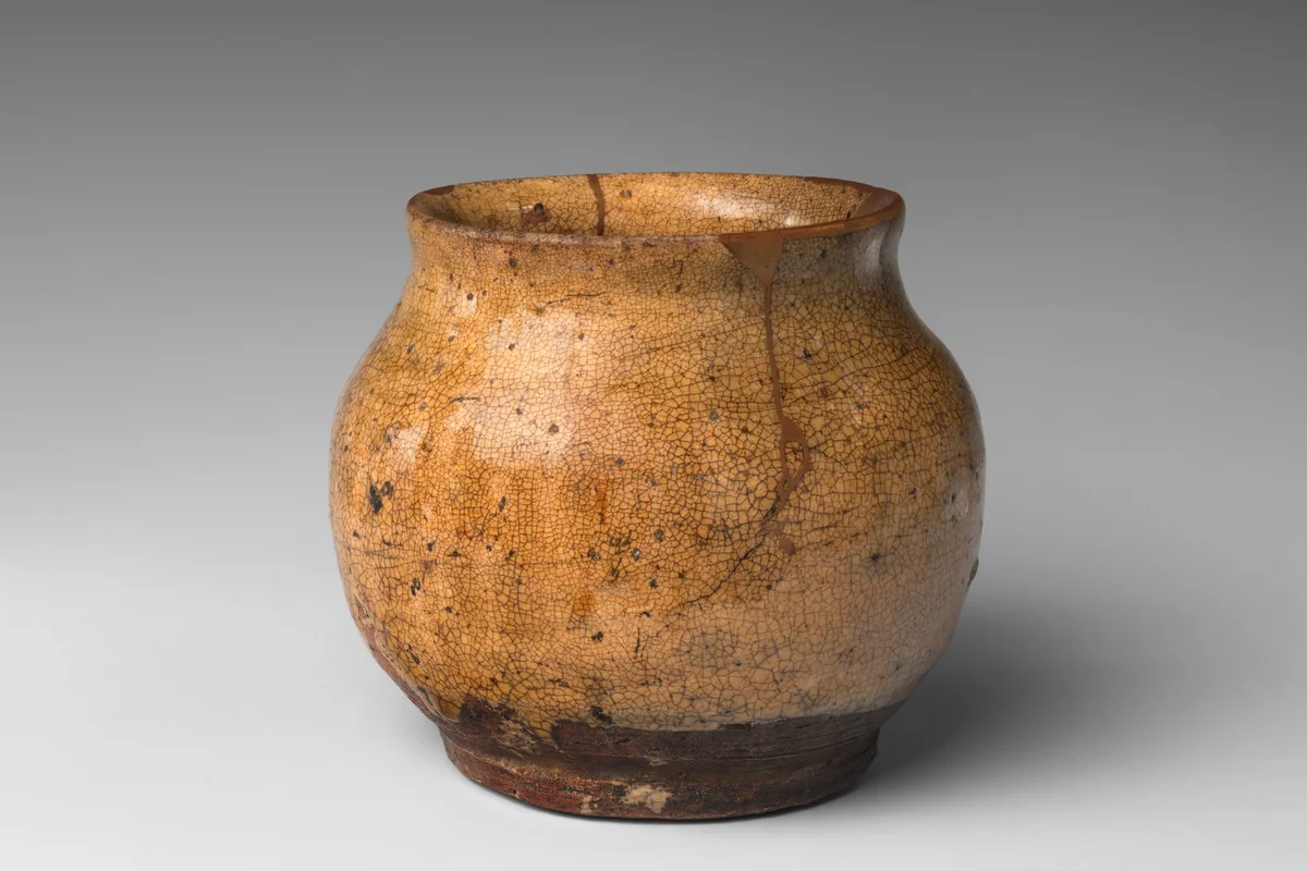A small rounded stoneware jar with a mottled amber ash glaze and a wide mouth, with gold lacquer filling cracks near the rim