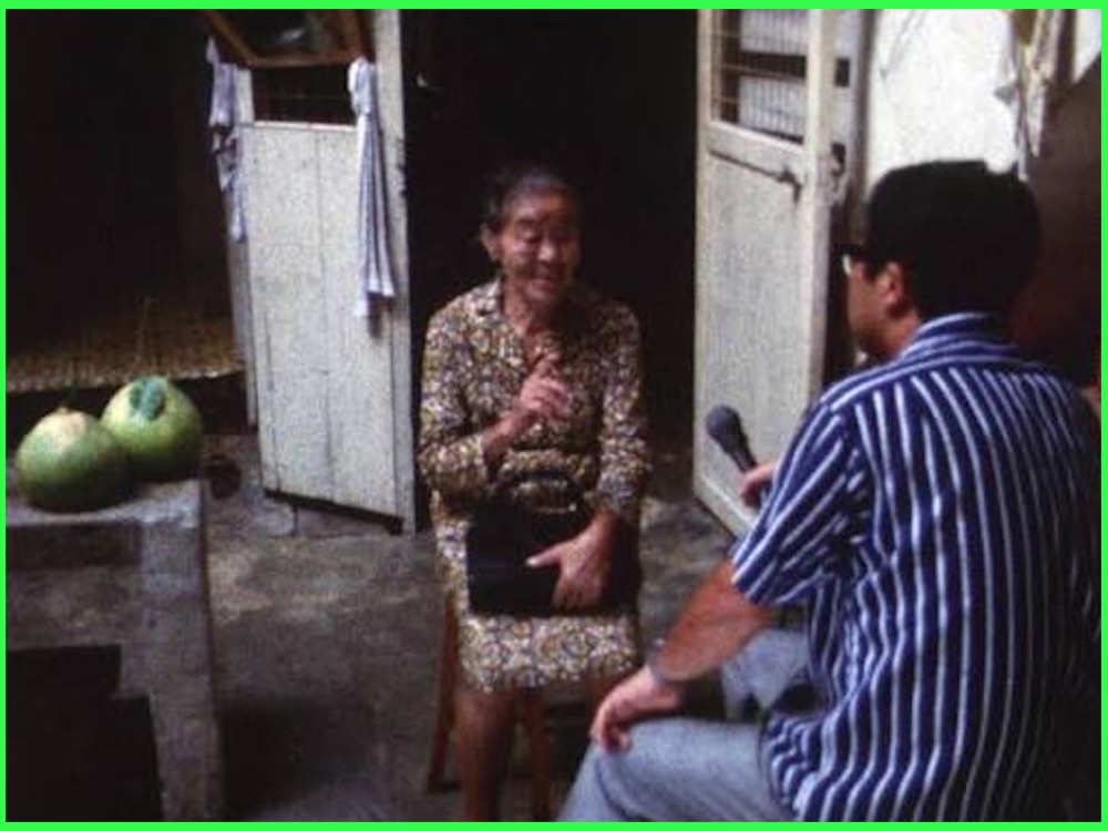 Colour still from Karayuki-san: The Making of a Prostitute (1975): an elderly woman in a patterned dress talks to an interviewer holding a microphone