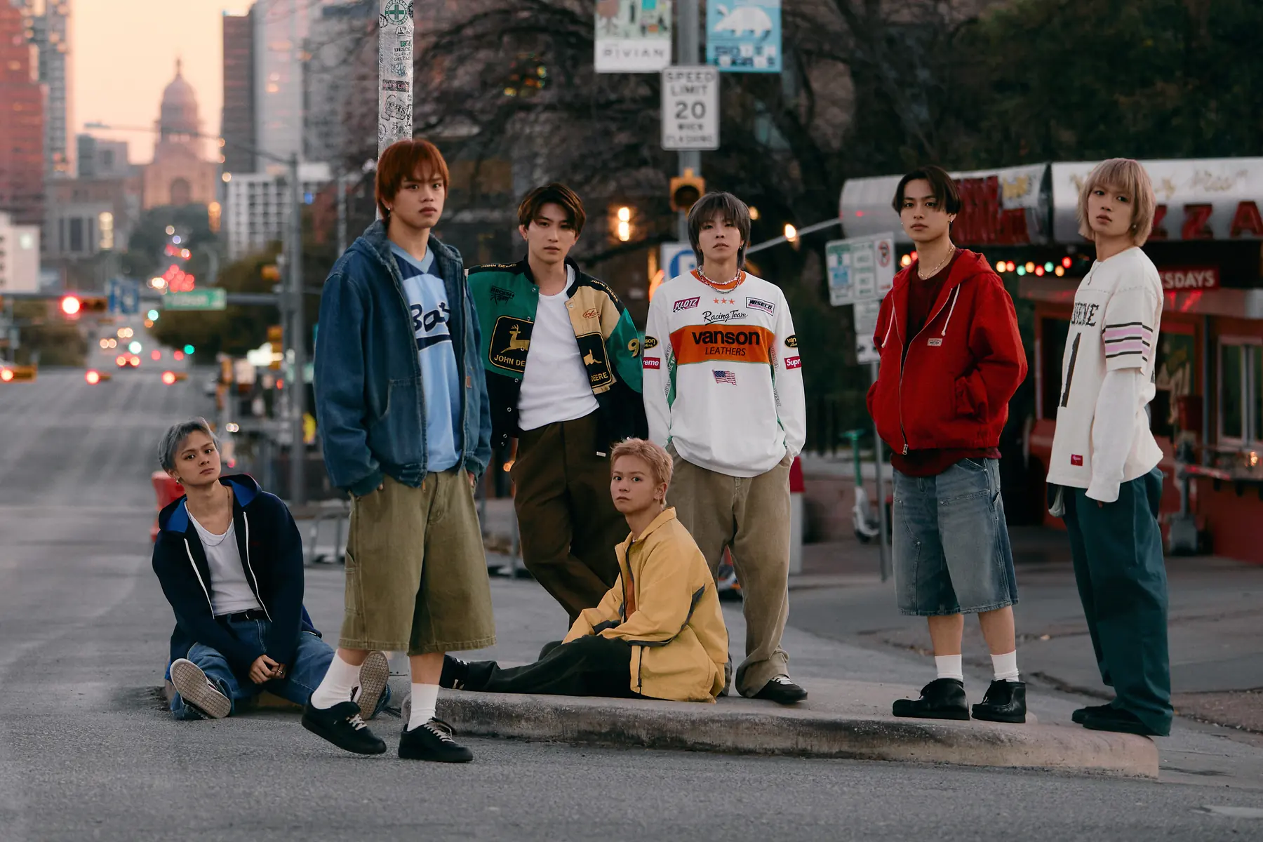 The seven members of KID PHENOMENON in streetwear on a kerb in Austin, Texas, at dusk, with the dome of the Texas State Capitol in the distance