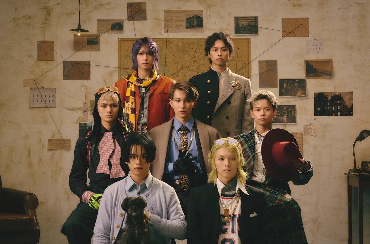 The seven members of KID PHENOMENON in vintage suits, scarves and hats, posed in front of a wall of maps and newspaper cuttings joined by red string, with one member holding a small dog in the front row