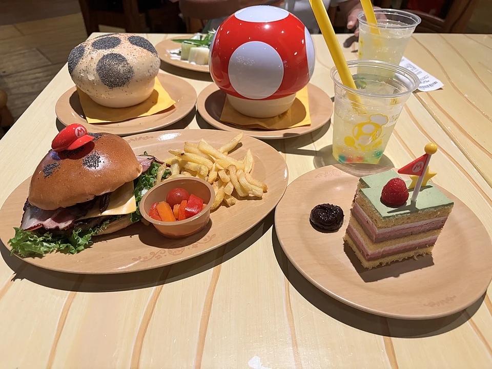 Lunch at Kinopio’s Cafe in Super Nintendo World: a burger with a Mario cap pick, the Super Mushroom Pizza Bowl, a layered cake with a goal flag and lemon drinks
