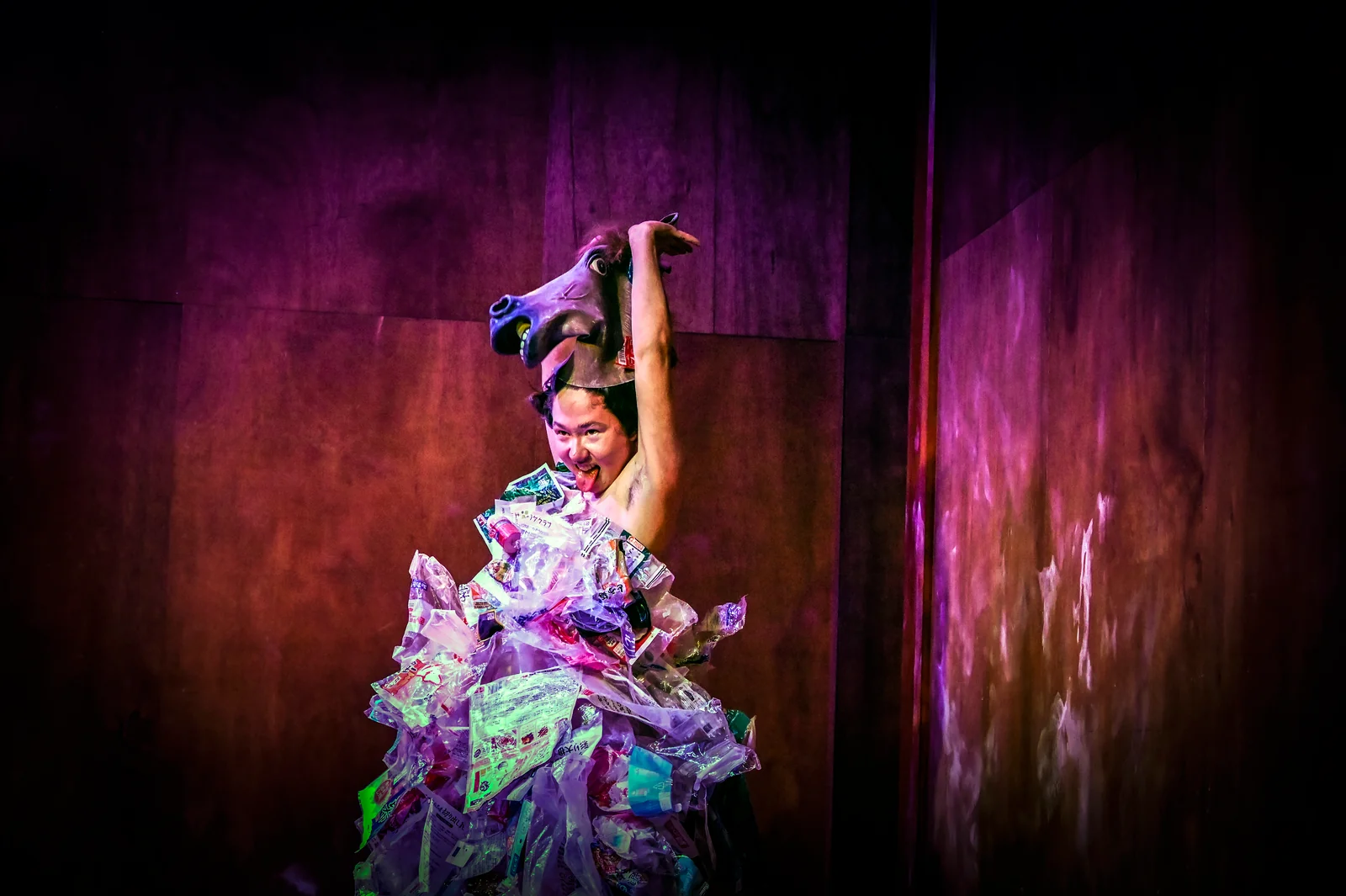 Mai Burns in a dress of layered clear plastic raises an animal mask above her head against a purple lit wooden wall