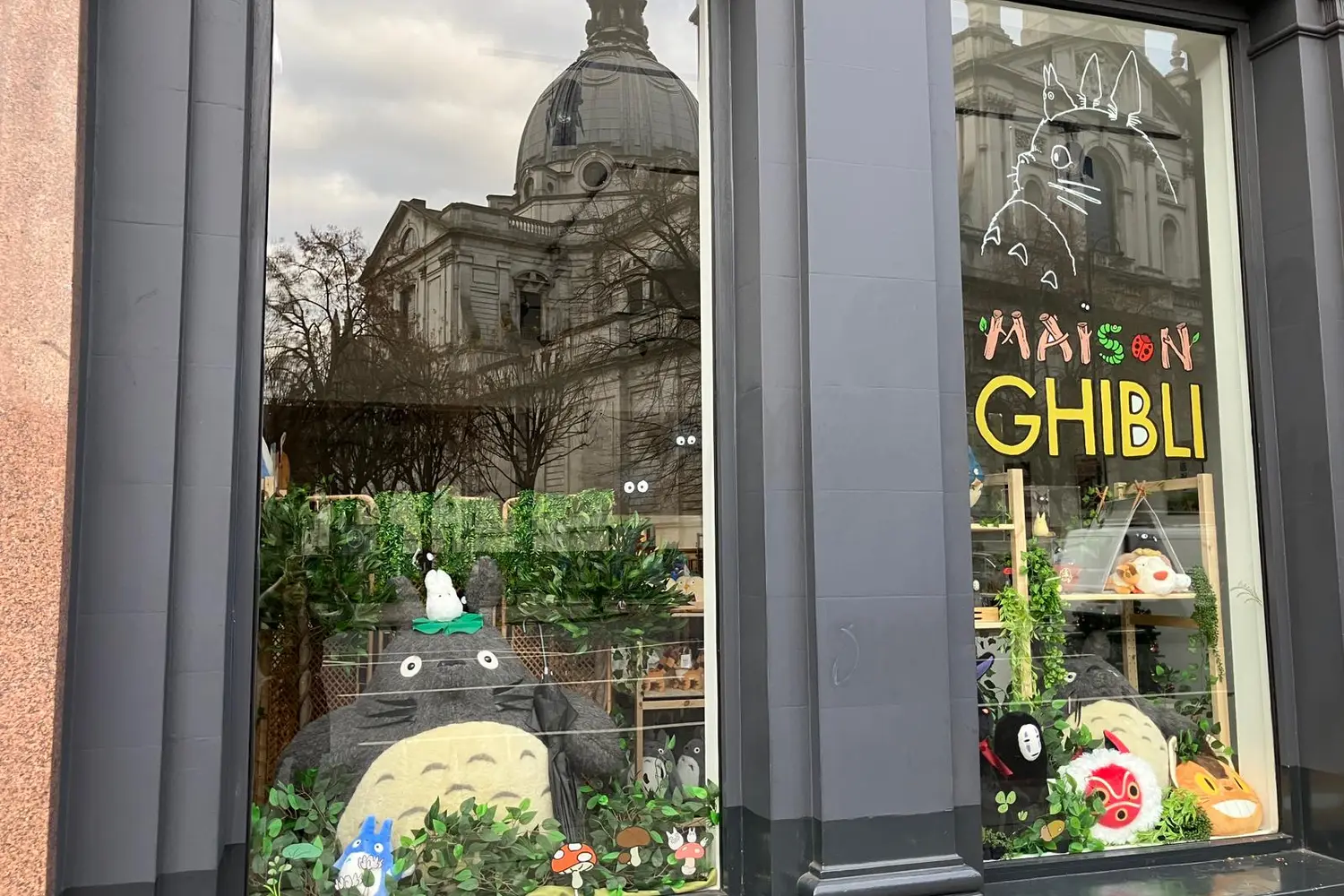 Maison Ghibli shop window with a giant Totoro plush among leaves, the Brompton Oratory dome reflected in the glass