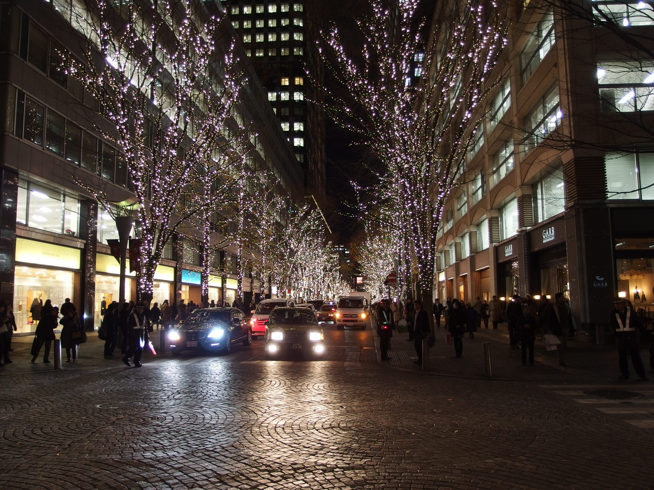 Trees wrapped in white fairy lights along a cobbled street in Marunouchi, Tokyo, at night, with office buildings and passing cars