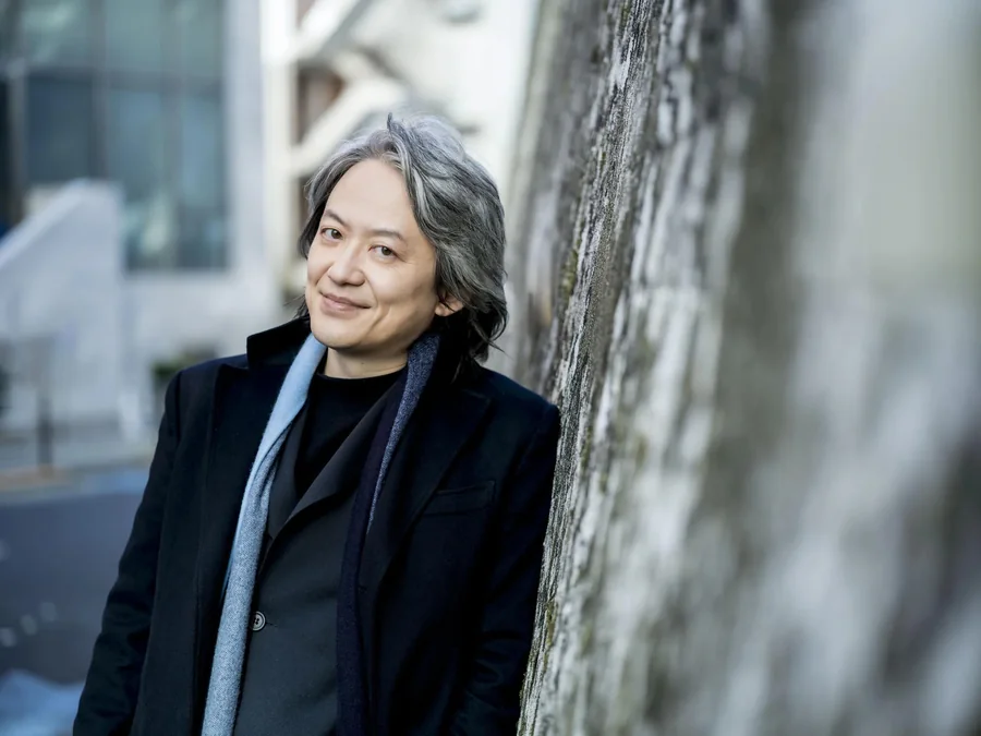Conductor Masato Suzuki, principal conductor of Bach Collegium Japan, smiling and leaning against a wall in a dark coat and blue scarf