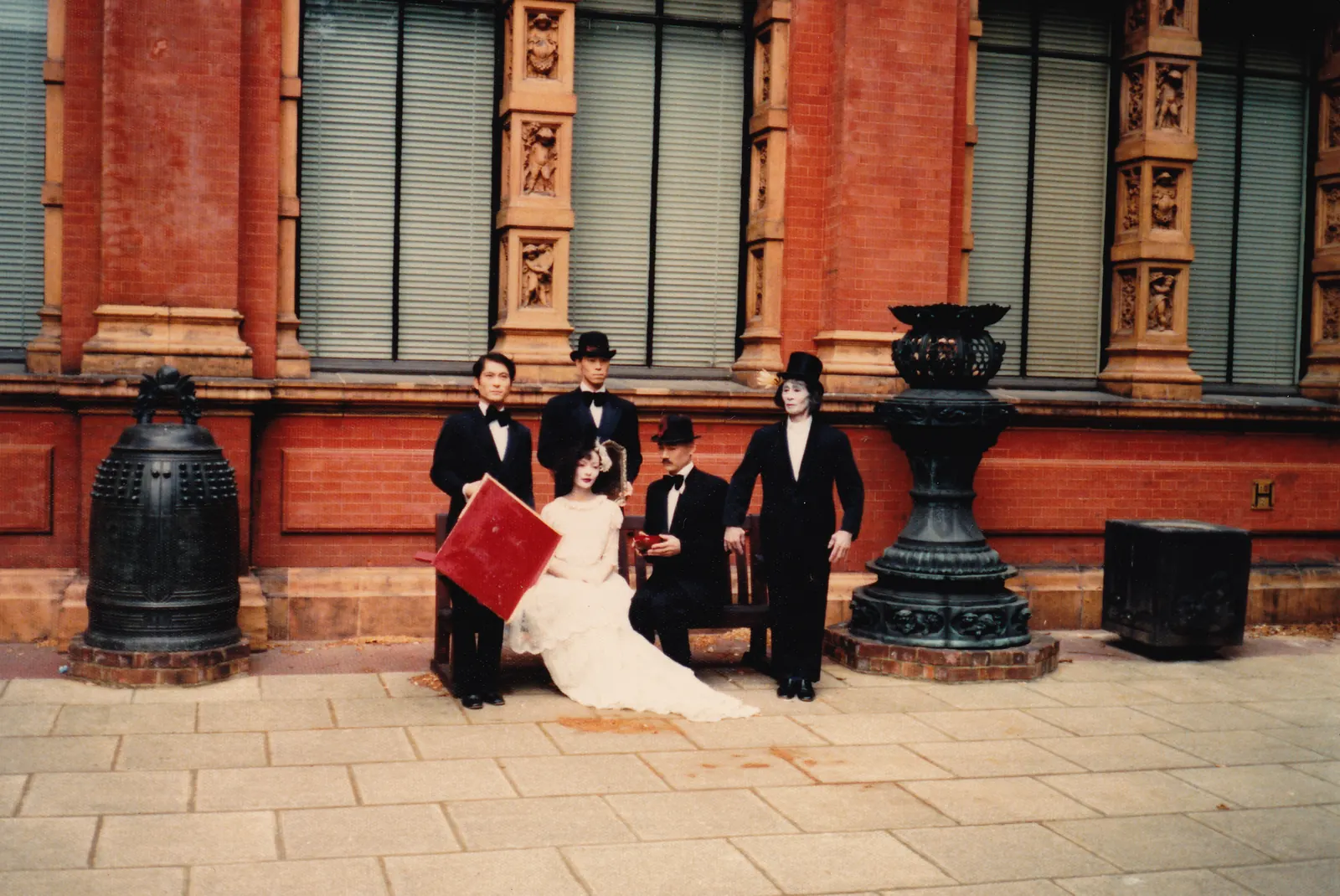Kazuo Ohno in a top hat stands with four members of the Ozen cast outside the red brick V&A in London, Mitsuyo Uesugi seated on a bench in a white gown