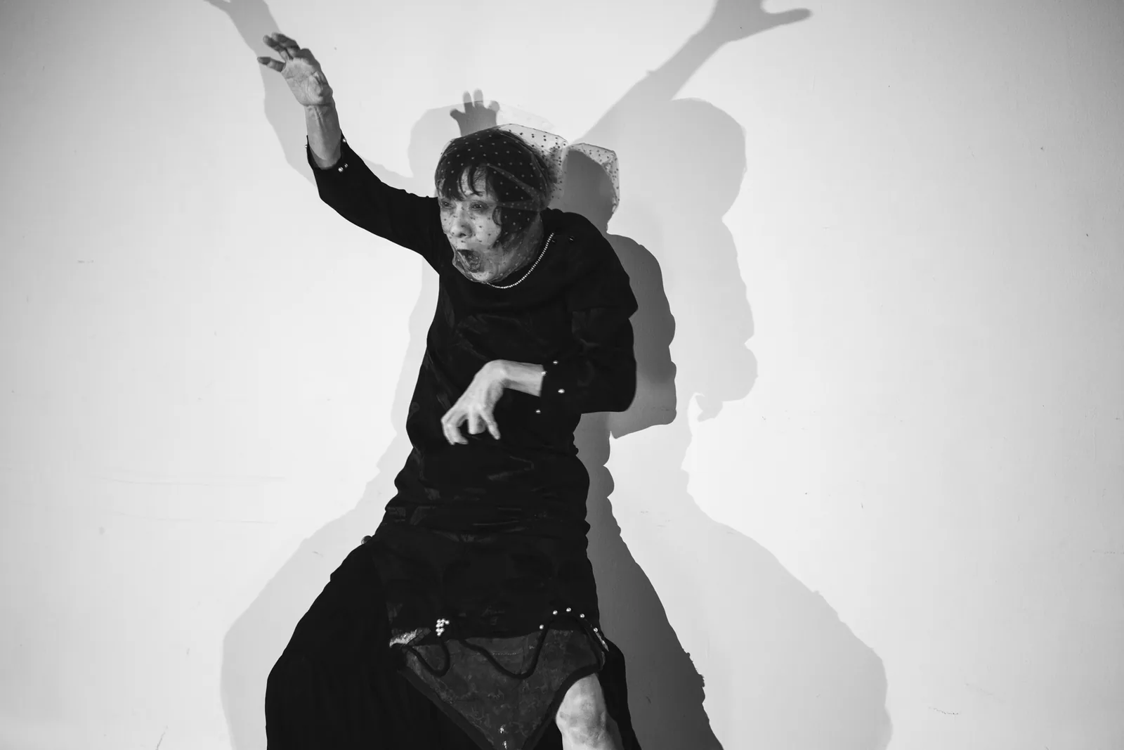 Mitsuyo Uesugi in a black dress and dotted veil throws one arm up against a white wall, mouth open, her shadow spread behind her