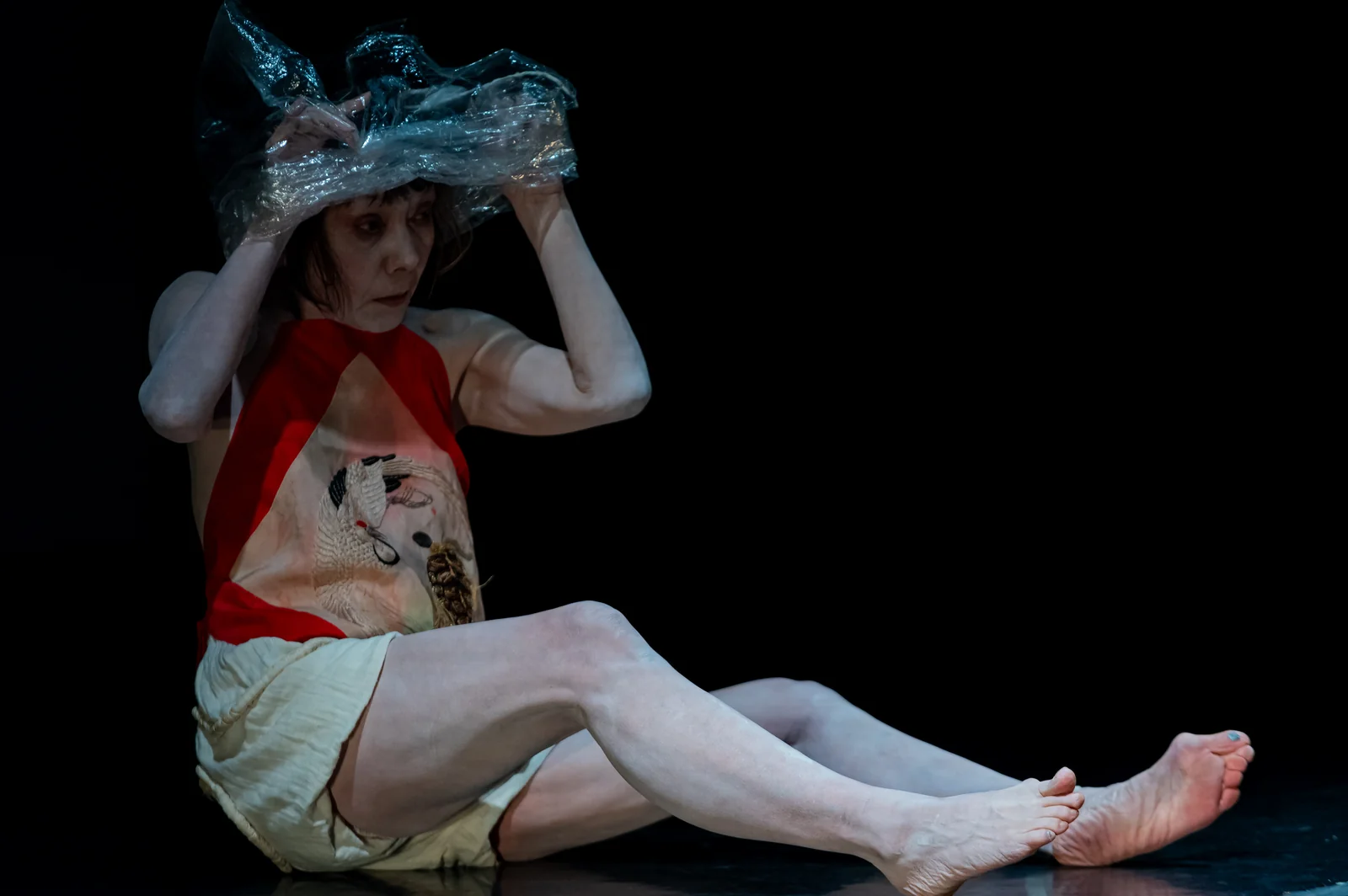 Mitsuyo Uesugi sits on a black stage with her legs stretched out, holding a sheet of clear crumpled plastic over her head
