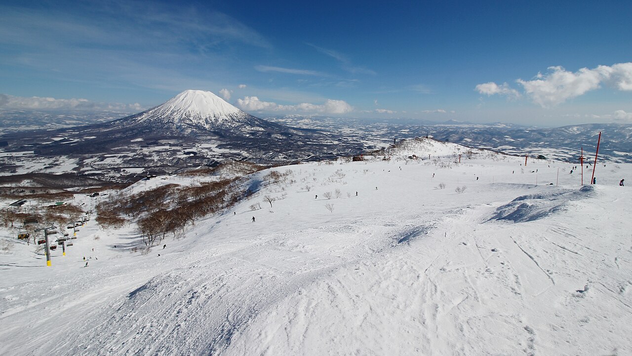 Mount Yotei under a blue sky seen from the snowy upper slopes of Niseko Annupuri, with skiers and a chairlift below