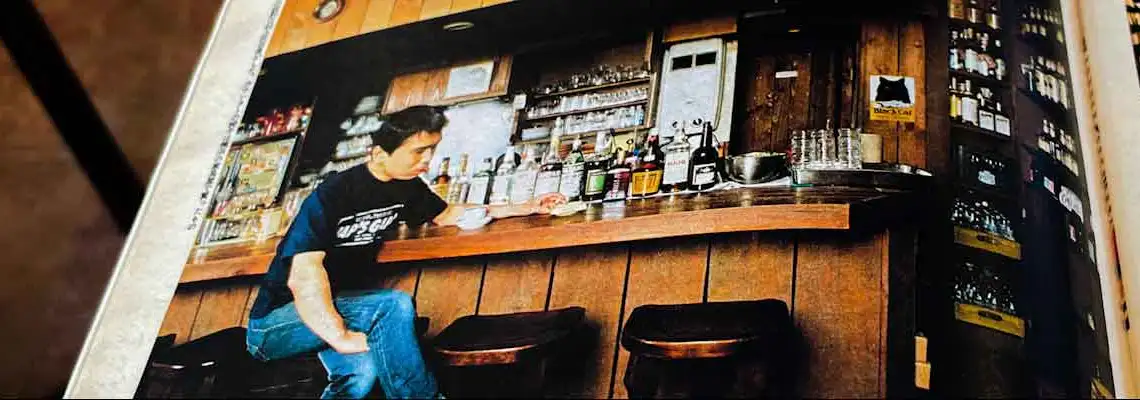 Haruki Murakami sitting on a stool at the wooden counter of his jazz bar Peter Cat, with bottles and glasses lined up behind the bar