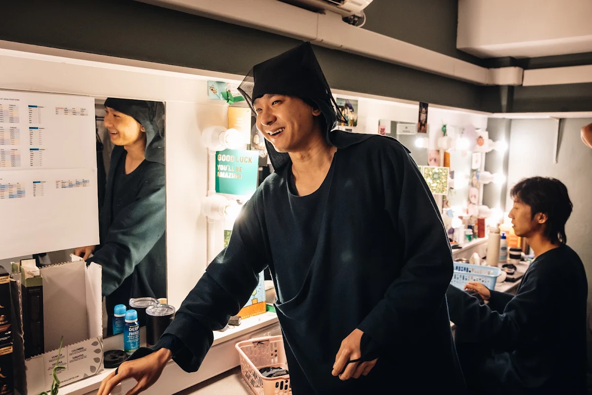 A puppeteer in a dark hood grinning in a dressing room, with colleagues at the mirrors behind