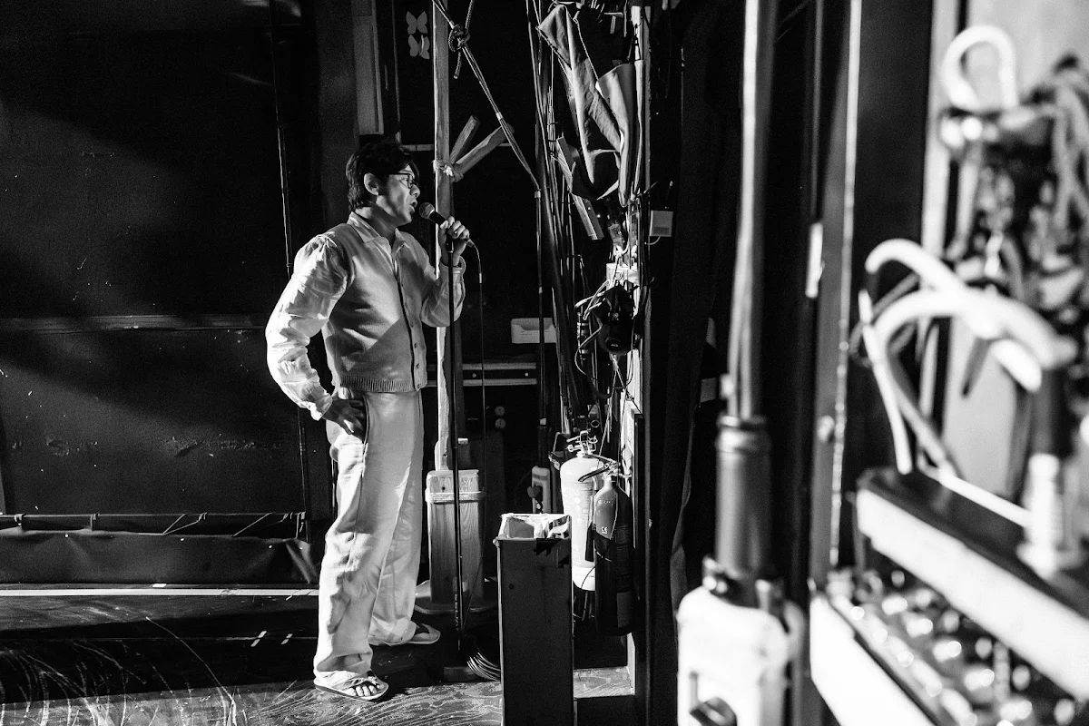 Dai Tabuchi in a light suit standing among the rigging and cables in the wings