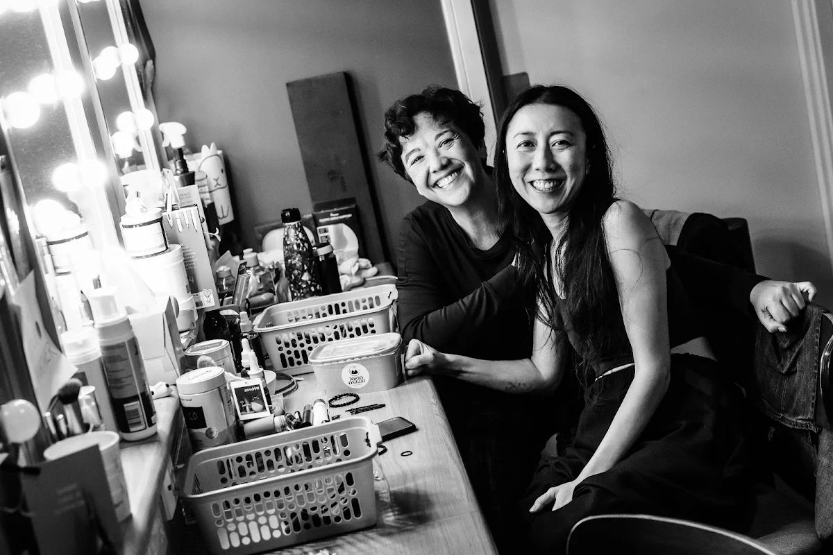 Jacqueline Tate and Phyllis Ho smiling at a dressing room mirror lined with bulbs and make up
