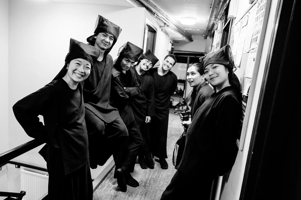 Black and white photograph of seven puppeteers in dark robes and hoods lined up along a backstage corridor