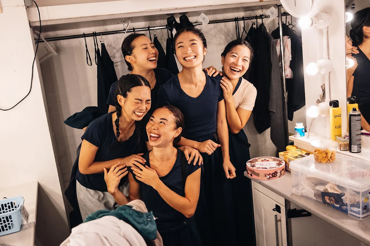 Five performers in dark costumes laughing and hugging in a dressing room